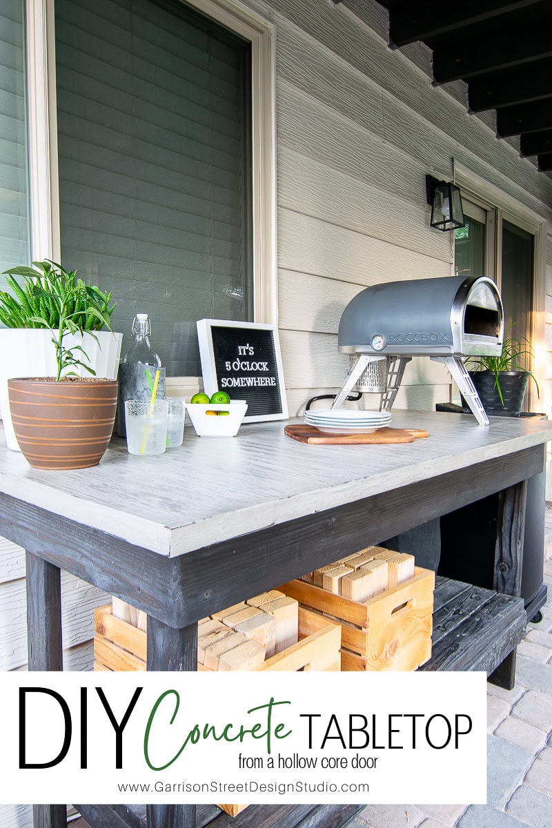 Outdoor table with a concrete tabletop crafted from a hollow core door, adorned with potted plants, glasses, a chalkboard sign, and a pizza oven. Wooden crates nestle beneath. Text on image says, "DIY Concrete Outdoor Table."