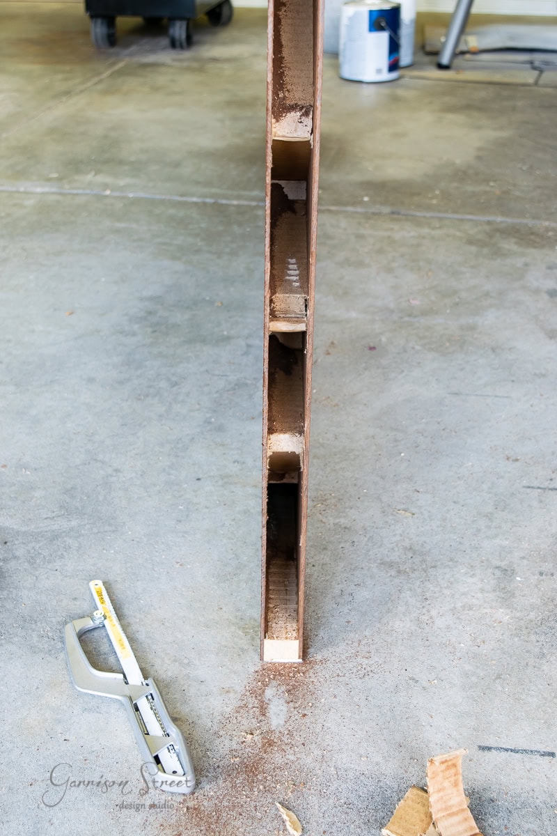 A close-up of the cut edge of a hollow core door reveals its cardboard supports. Positioned upright on a concrete floor, amid wood shavings, with a saw and paint cans nearby.