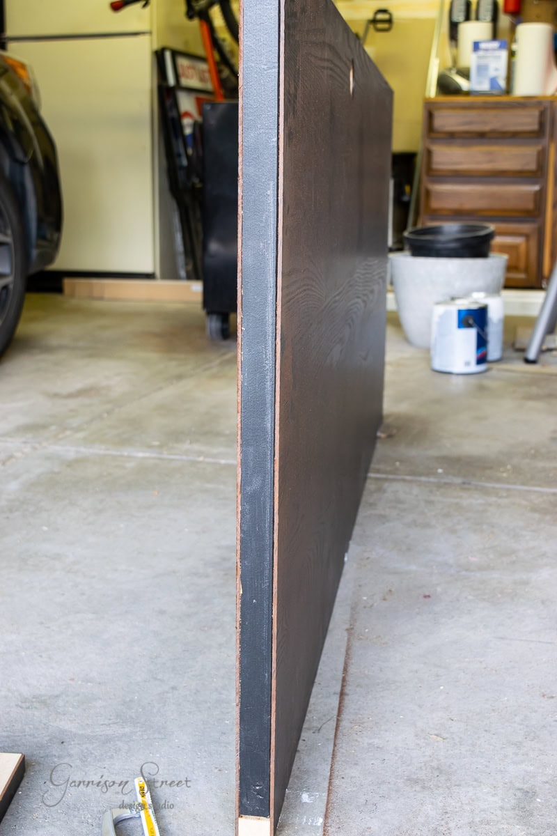 A black wooden door panel leans against a garage workbench, surrounded by various tools and supplies like a paint can and toolbox.