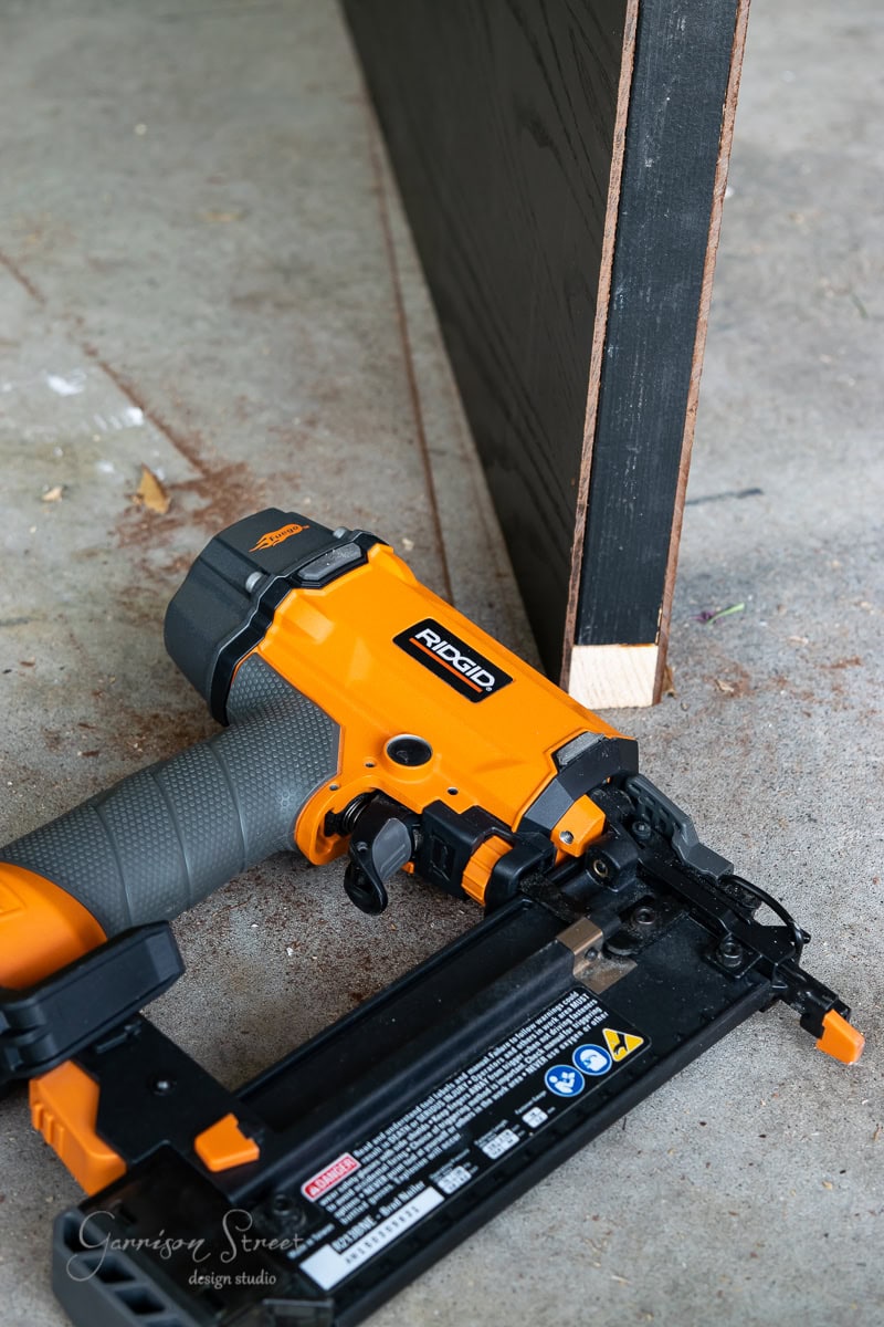 An orange nail gun is resting on a concrete surface, poised next to a piece of wood leaning against a wall. The wood boasts a white corner, and several labels are visible on the tool.