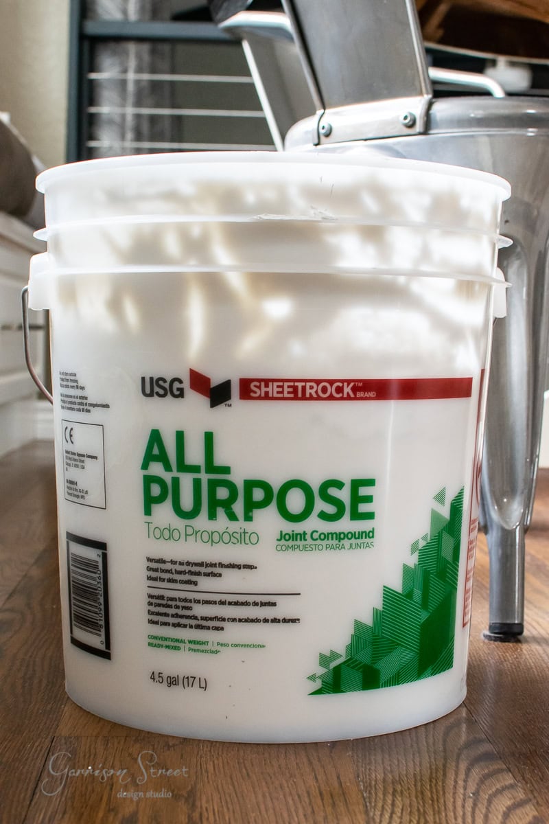 A large white bucket of Sheetrock brand All Purpose Joint Compound sits on a wooden floor, next to framed canvas art in the background. The label is printed in English and Spanish.