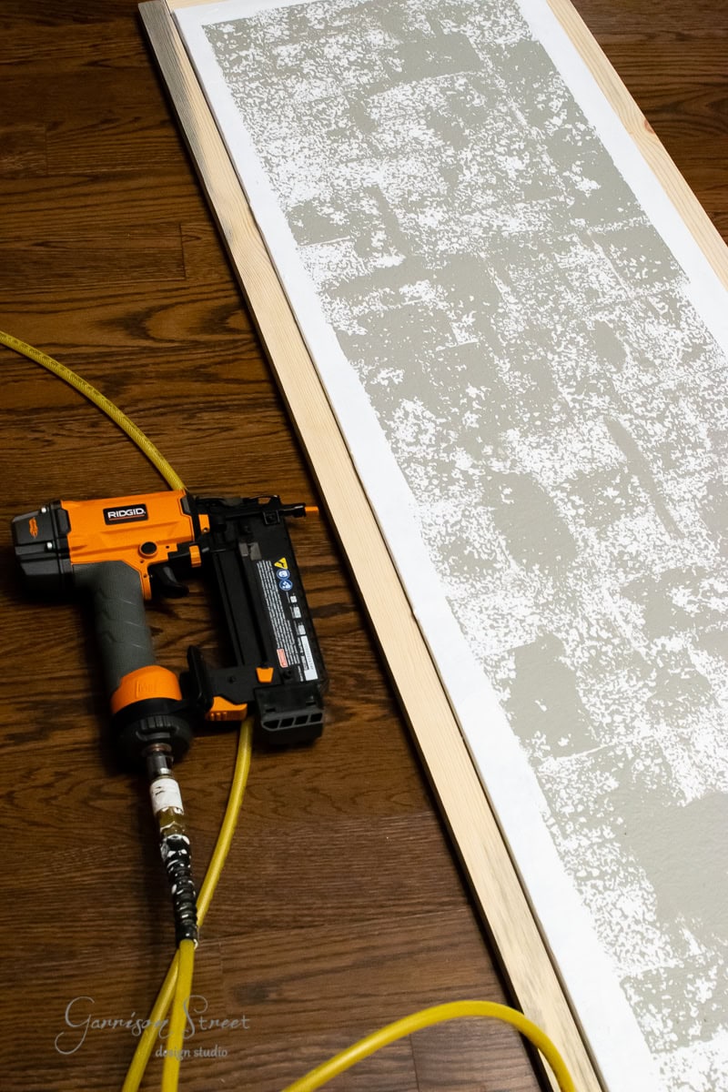 A nail gun with an orange and black design is connected to a yellow air hose, resting on a wooden floor. Beside it lies a piece of plywood, its textured surface reminiscent of gray painted framed canvas art.