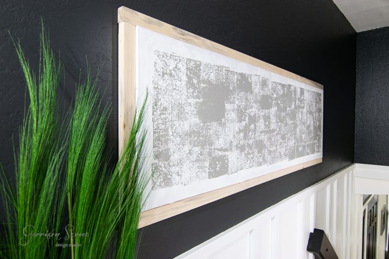 A modern framed canvas art piece featuring textured gray patterns on a white background is set in light wood. It elegantly hangs against a dark wall above white paneling, while tall green grass-like plants add a touch of nature to the scene from the foreground on the left side.