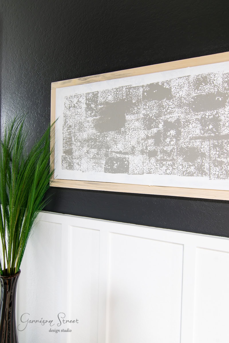 A minimalist framed canvas art piece with a textured gray brick pattern is mounted on a black wall. Below it, white paneling adds contrast. A green plant in a vase stands to the left, providing a touch of nature.