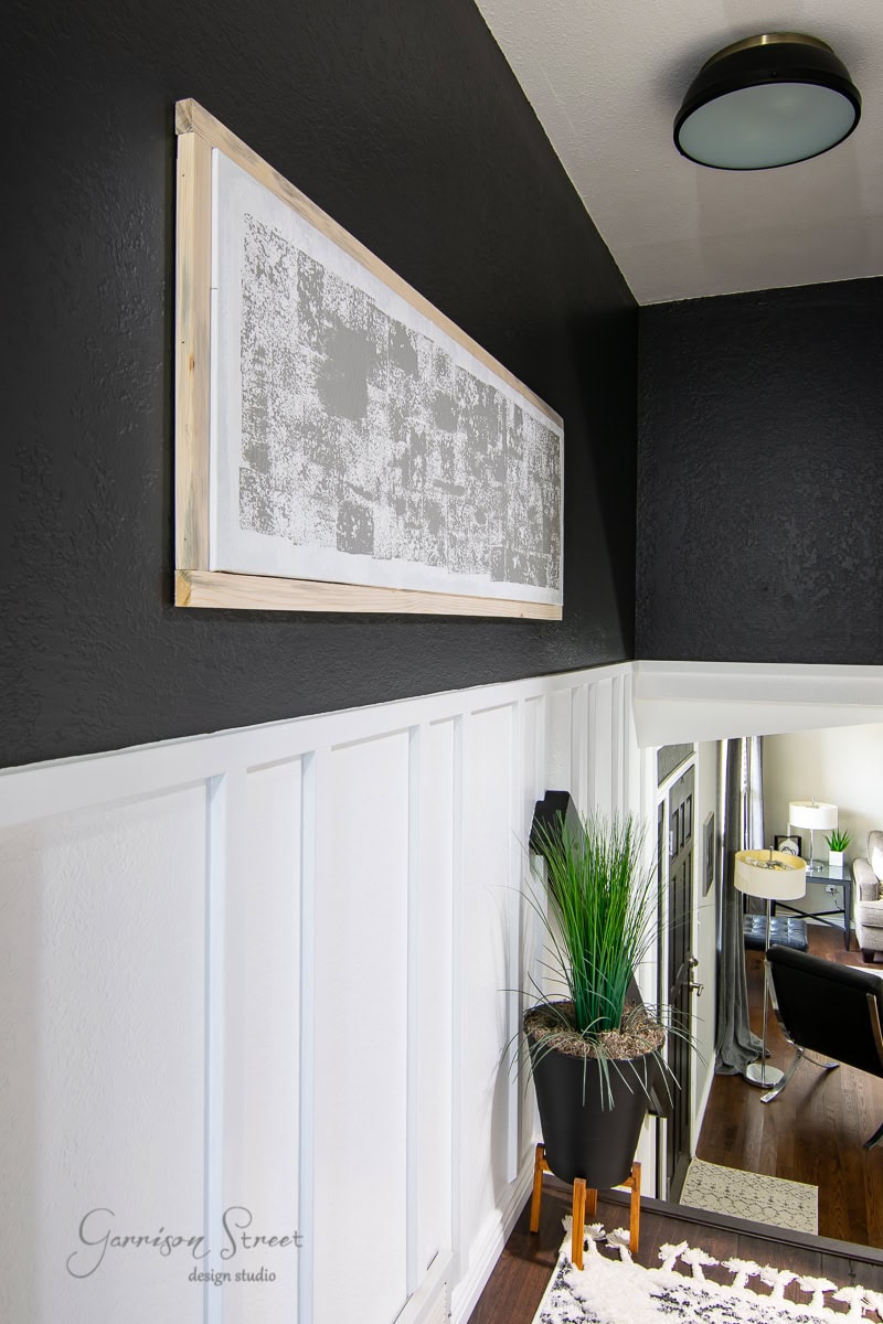 A modern interior with black and white walls showcases a striking framed canvas art piece. A tall green plant in a black pot sits on a stand nearby, adding a touch of nature. In the background, hints of an inviting living area are visible, completing this harmonious space.