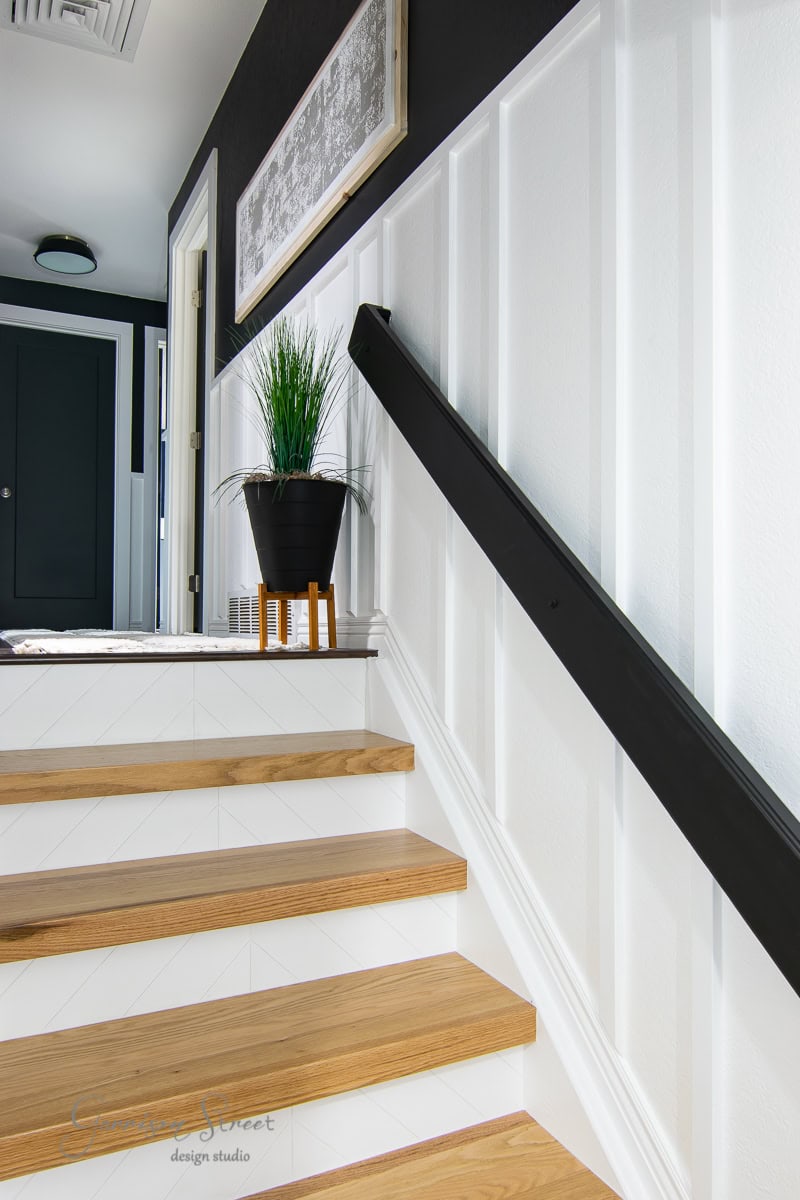 Modern staircase with light wood steps and white panel walls. A black handrail runs along the side, complemented by framed canvas art that adds a touch of elegance. A potted plant on a small stool decorates the landing, while a black door is visible at the top of the stairs.