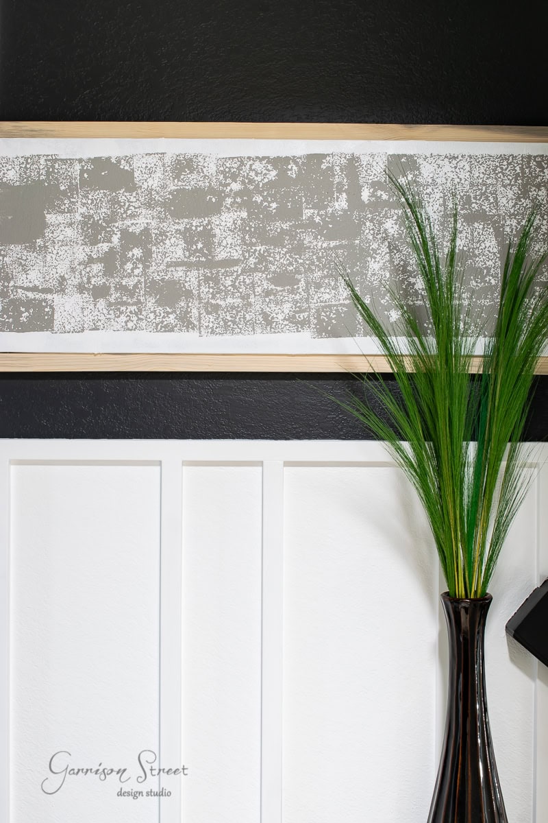 A minimalist interior design boasts a dark vase with tall green grass-like plants against a black and white paneled wall. Above, framed canvas art in shades of gray and white adds an abstract touch. The wall features the "Garrison Street design studio" watermark.