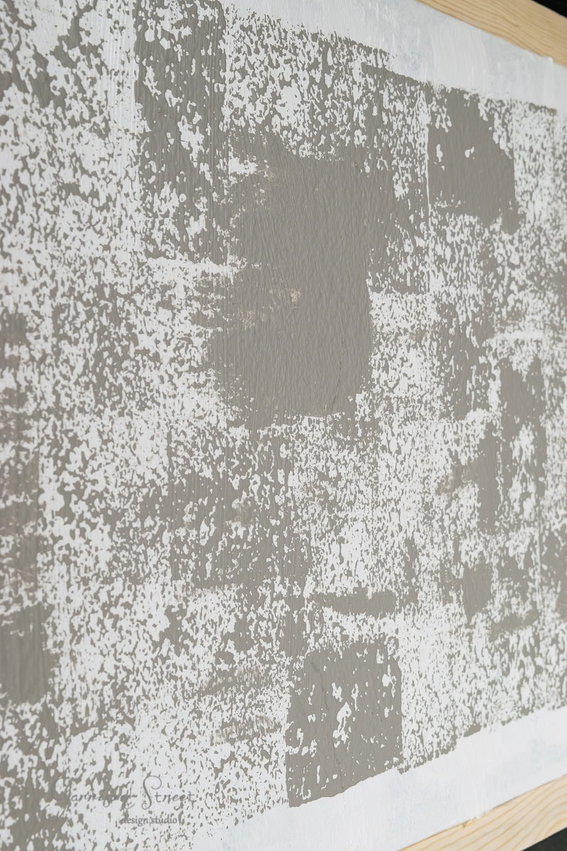 A close-up of a textured framed canvas art featuring a grid-like pattern of off-white and gray blocks. The design resembles a patchwork of varying shades and textures, creating an abstract, weathered appearance.