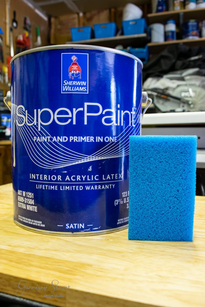 A blue can of Sherwin-Williams SuperPaint sits on a wooden table, accompanied by a blue sponge. In the background, slightly out of focus, shelves display various items, including framed canvas art that adds an artistic touch to the scene.