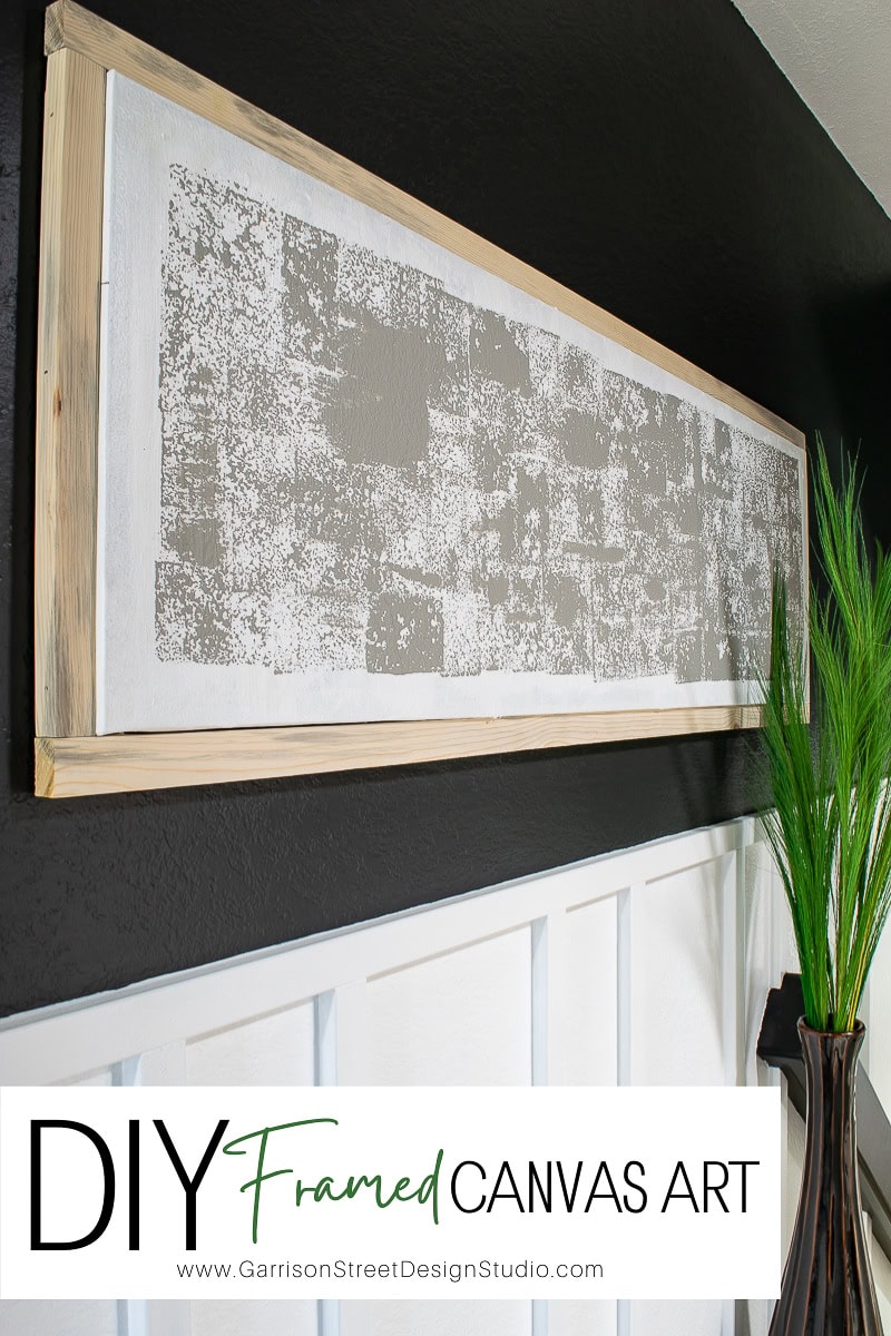 A framed DIY canvas art piece hangs on a dark wall above white wainscoting. The framed canvas features a textured, abstract gray and white design. A green plant is partially visible in the foreground.
