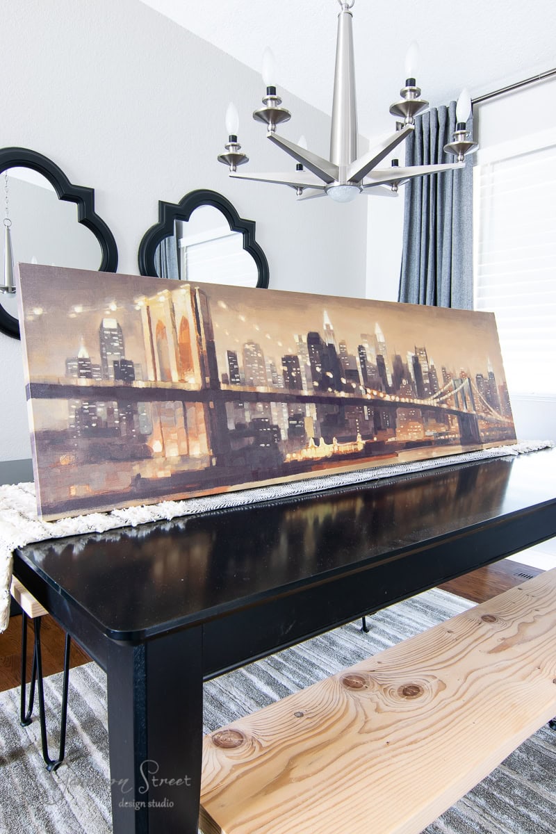 A large framed canvas art of a city skyline with bridges graces a wooden table in the dining room. The room features a modern chandelier, a black bench, and decorative wall mirrors, creating an elegant and contemporary atmosphere.