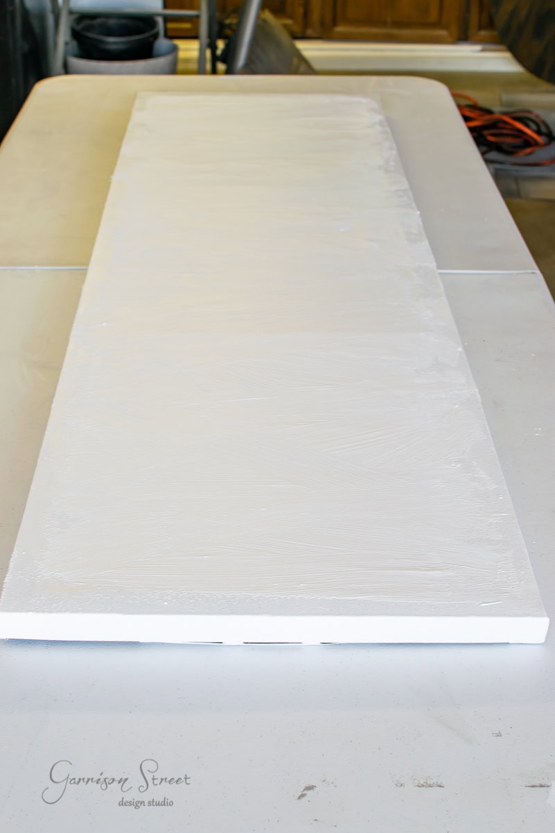 A long, rectangular wooden board on a table, coated with a layer of white primer, resembles a framed canvas art piece. The surface appears smooth and ready for painting. In the background, various tools and equipment are visible in a workshop setting.