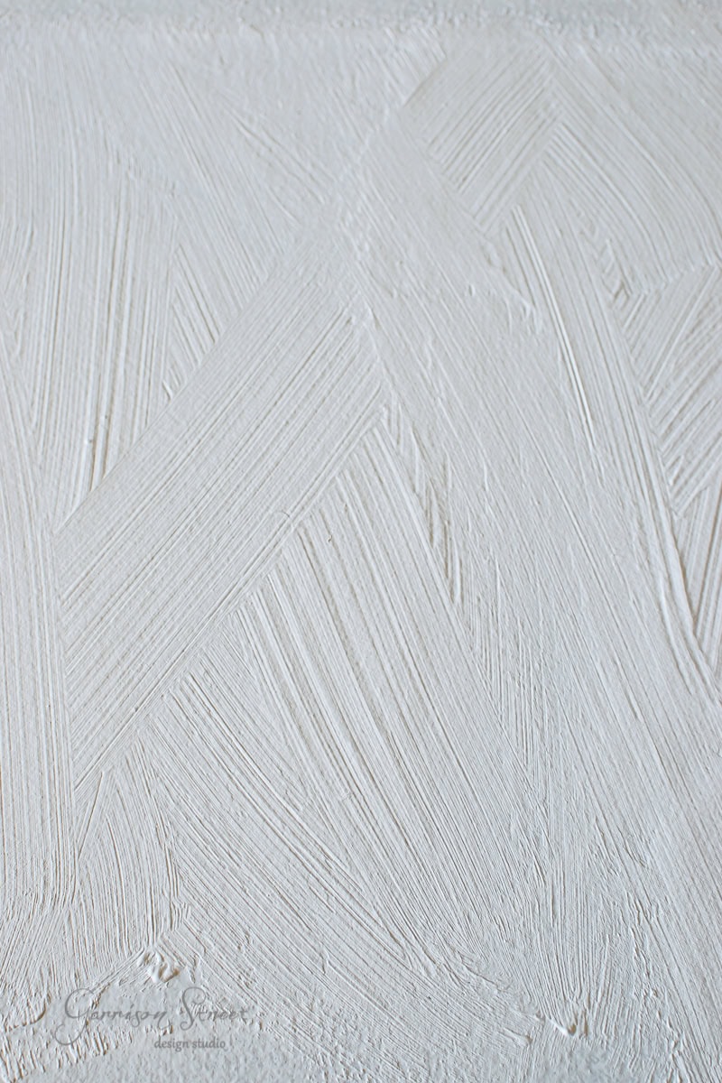 Textured white wall with visible brush strokes forming various intersecting diagonal patterns. The matte finish creates a subtle play of light and shadow, reminiscent of framed canvas art in its intricate detailing.