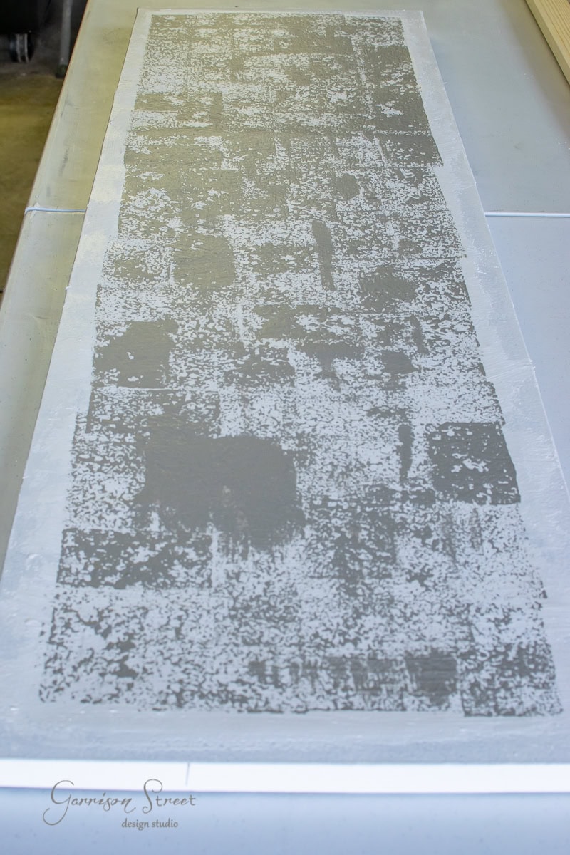 A textured, rectangular tabletop in progress, painted in a distressed gray and white pattern resembling framed canvas art, rests on a smooth surface. The logo "Garrison Street Design Studio" is faintly visible in the corner.