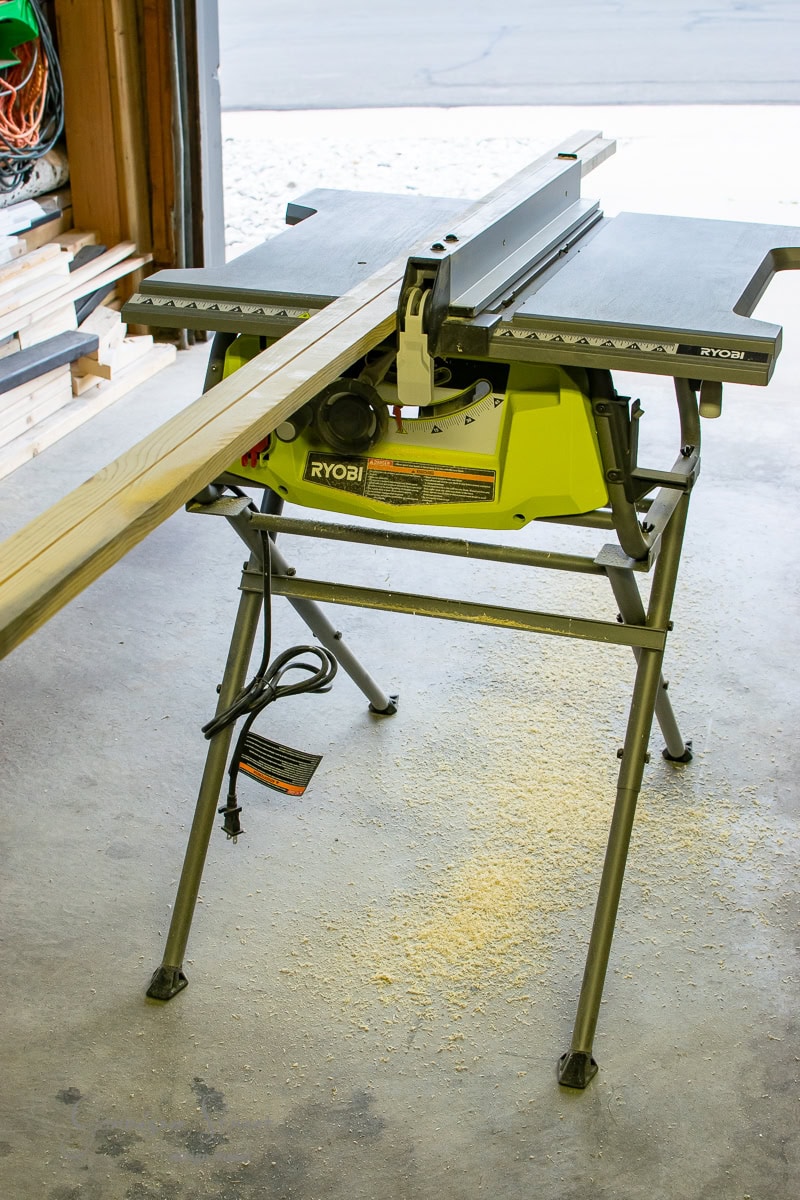 A Ryobi table saw hums in a garage, effortlessly slicing through a long wooden plank. Sawdust is scattered like fine art on the concrete floor around the setup. Shelves in the background hold additional wood pieces, echoing the precision of framed canvas art.