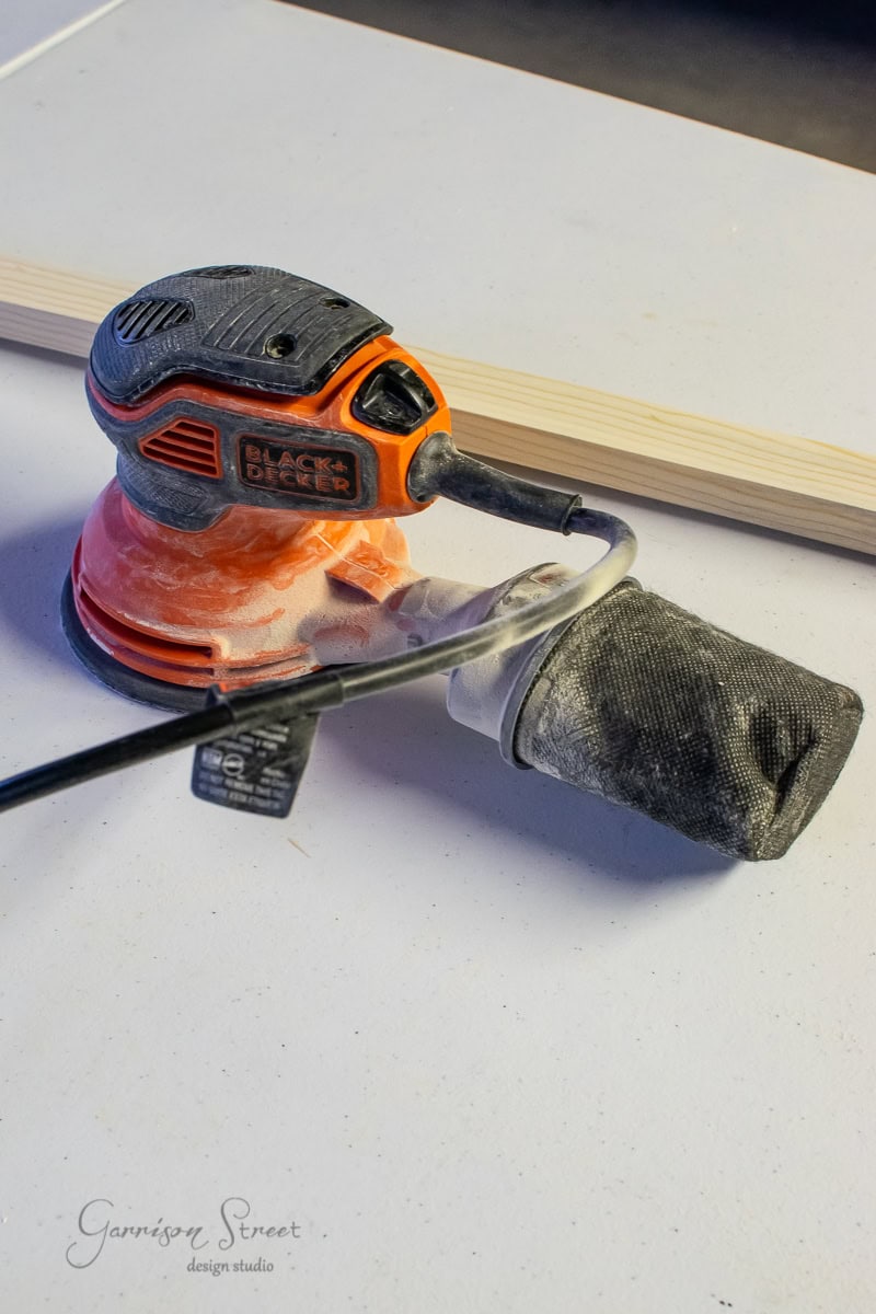 An orange and black Black & Decker sander with a dust collection bag attached rests on a white surface beside a piece of light-colored wood, resembling the smooth texture of framed canvas art. Dust is visible on both the table and the sander.