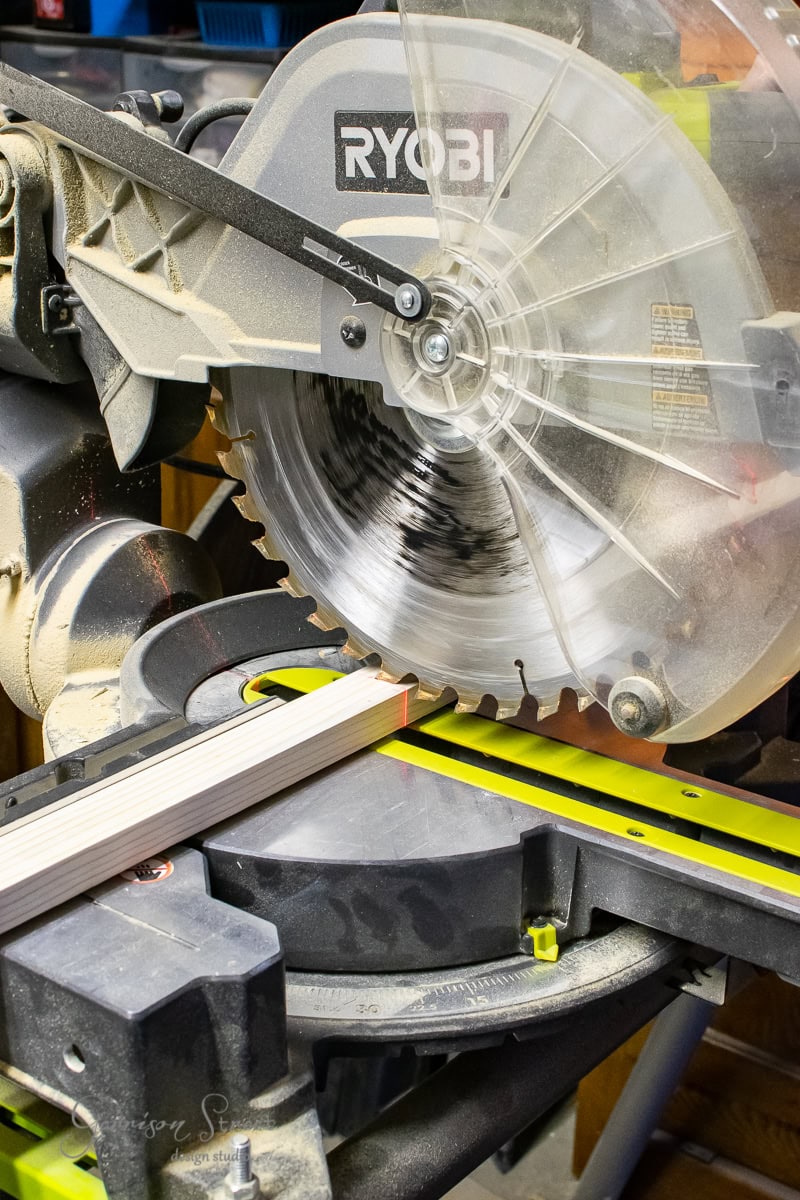 A power miter saw, bearing the Ryobi brand name, is actively cutting a piece of wood. The spinning blade is captured in motion, with sawdust accumulating around the tool like a framed canvas art in progress.