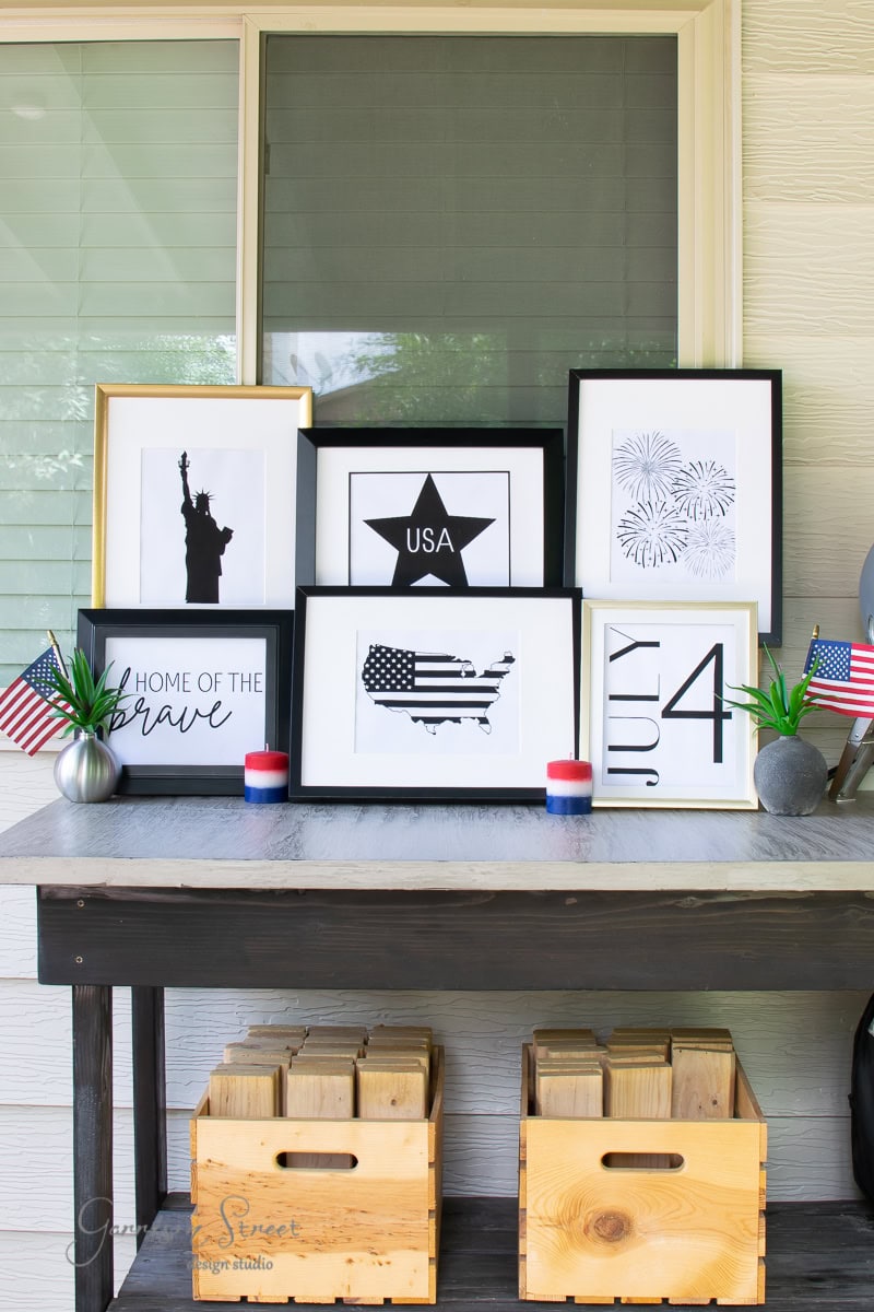 A decorative table showcases framed printable art with patriotic themes: a silhouette of the Statue of Liberty, an American flag map, and a star with "USA." Accentuated by text like "Home of the Brave" and "4th of July," small flags and candles complete the festive setup.