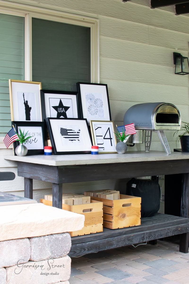 A patio table adorned with framed 4th of July-themed art, featuring the Statue of Liberty and an American flag design. Small American flags and two wooden crates decorate the table, while a small pizza oven sits to the right.