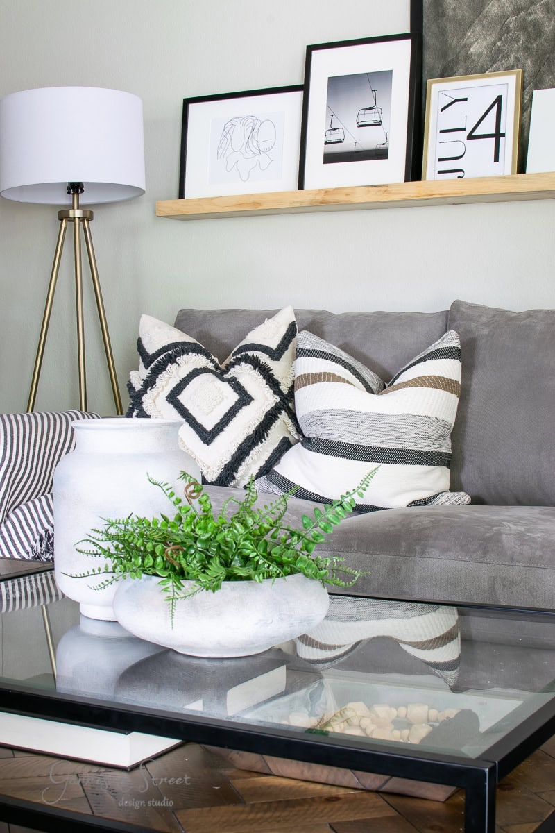 A modern living room features a gray sofa adorned with geometric and striped pillows. A glass coffee table holds a potted fern and decorative items. A floor lamp stands nearby, and printable art is displayed on a wall shelf behind the sofa.