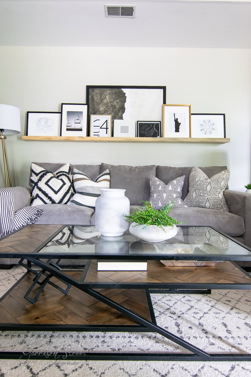A modern living room with a gray couch adorned with patterned cushions. A glass coffee table sits on a geometric rug. A shelf behind the couch displays framed art and printable photographs. A floor lamp is visible on the left.