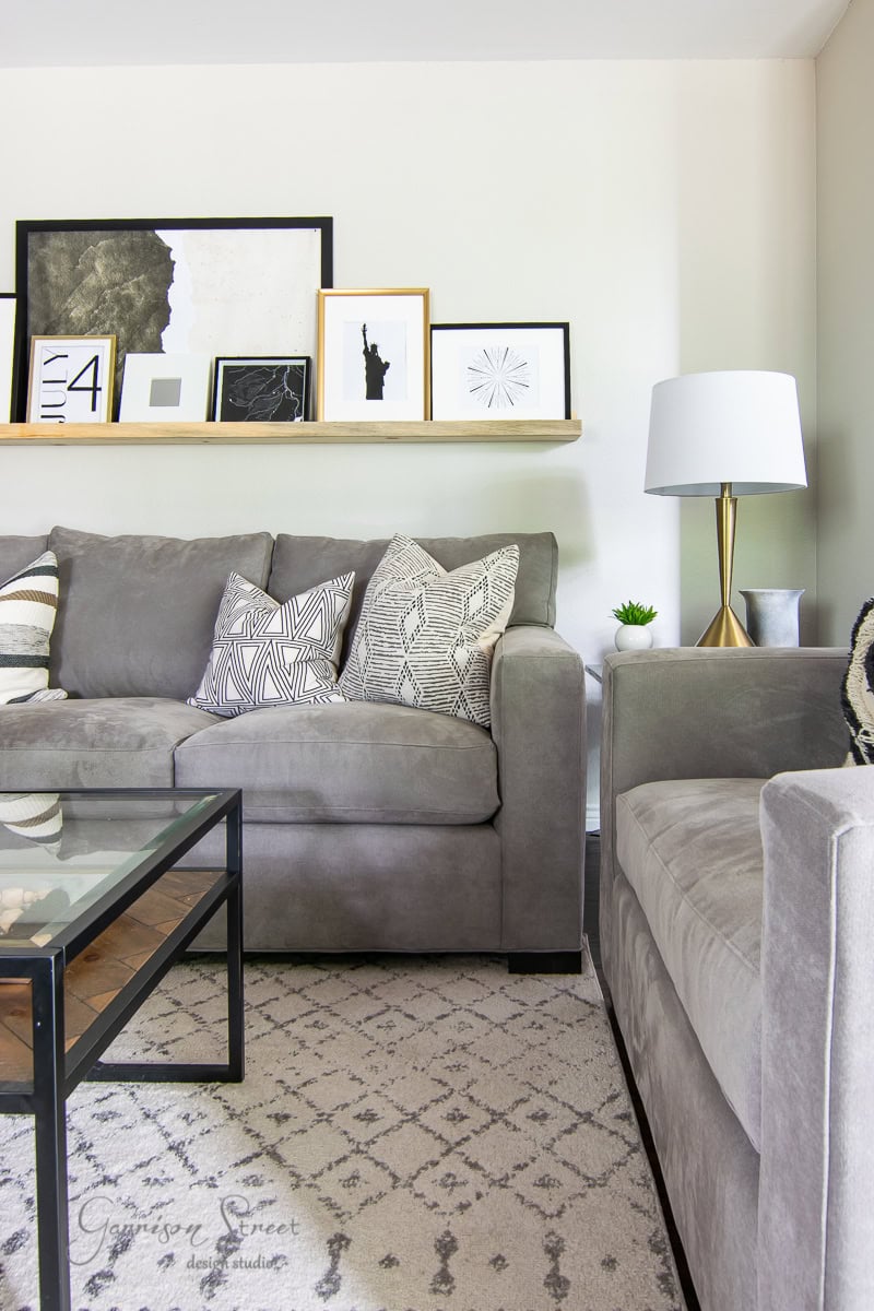 A modern living room features a gray sofa and armchair adorned with patterned cushions, complemented by a glass coffee table and geometric rug. A shelf displays framed, printable art pieces. A table lamp with a white shade sits gracefully on the side table.