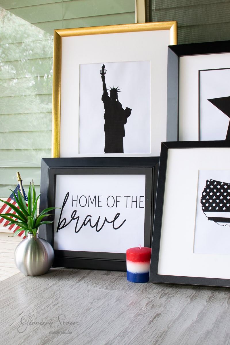 Framed artwork arranged on a table features a "Home of the Brave" printable art, Statue of Liberty silhouette, and star pattern with an American flag theme. A small American flag and a red, white, and blue candle complete the patriotic display.