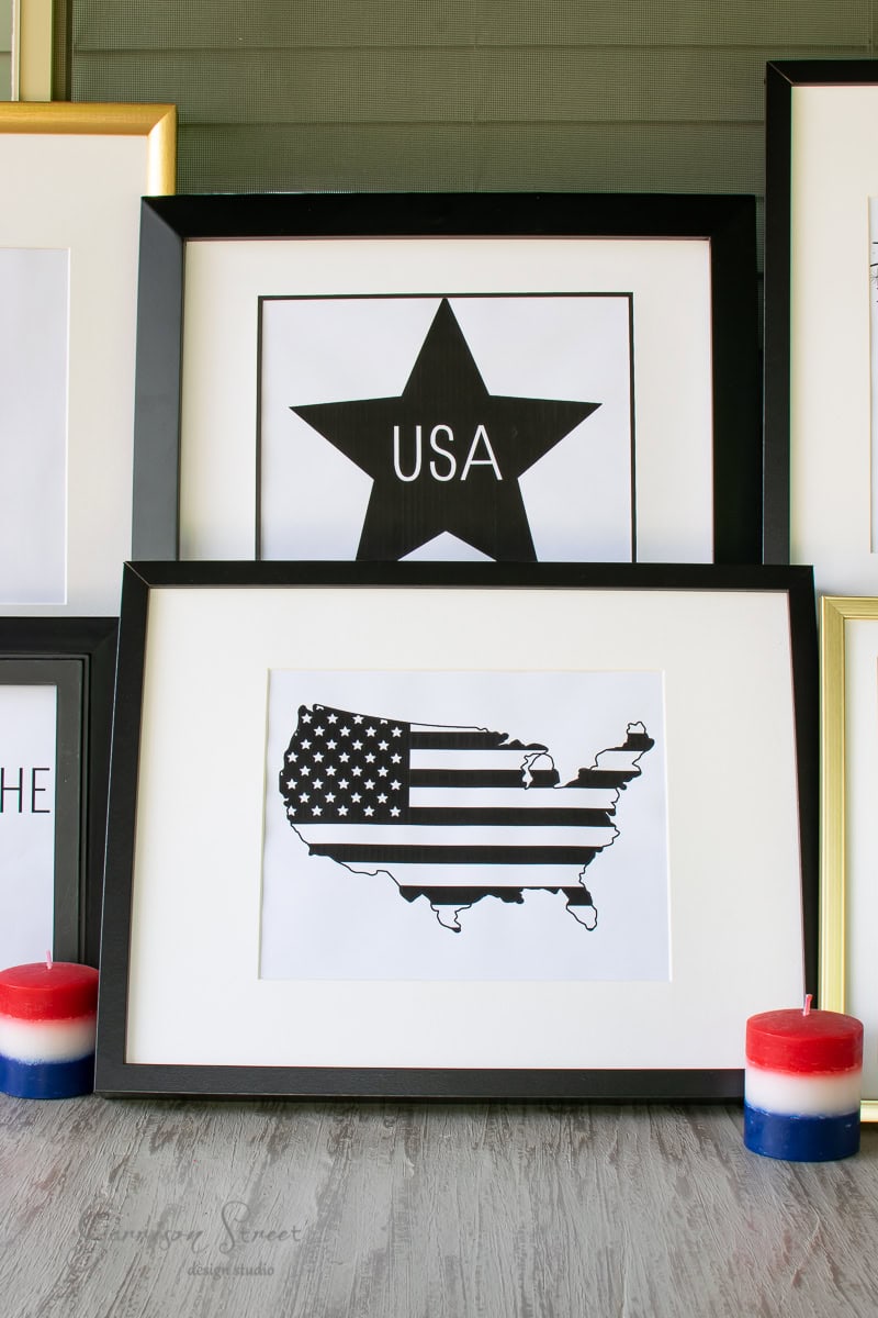 Framed artwork on the floor showcases a striking black and white American flag in the shape of the United States. Behind it, a framed printable art piece features a star with "USA" inside. Two red, white, and blue candles flank the setup.