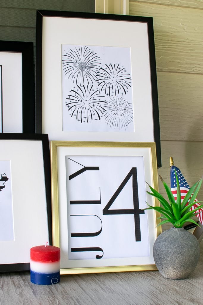Home Framed printable art with "4 July" typography and black fireworks illustrations adorns the wall. A small American flag and a red, white, and blue candle sit on the table beside a potted plant, celebrating the spirit of 4th of July.