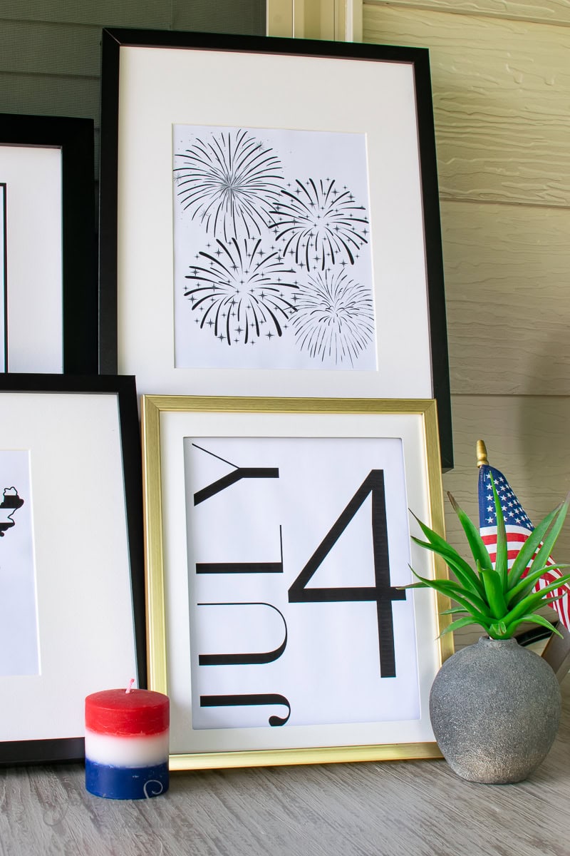 Framed printable art with "4 July" typography and black fireworks illustrations adorns the wall. A small American flag and a red, white, and blue candle sit on the table beside a potted plant.