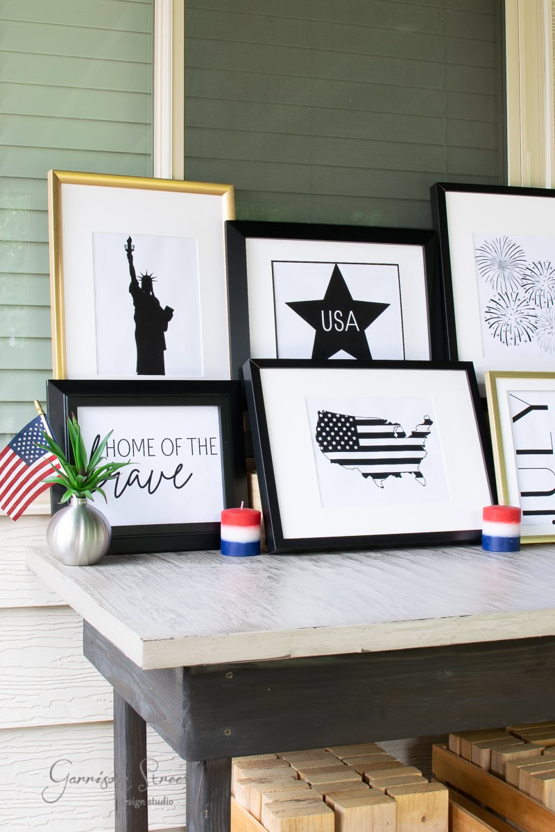 A table displays framed patriotic art, featuring a silhouette of the Statue of Liberty, a star with "USA," and a map with stripes. An American flag graces a vase alongside red, white, and blue candles.