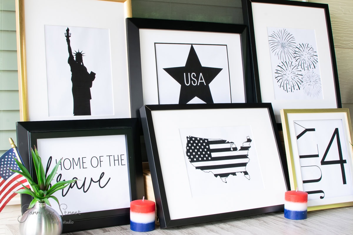 A collection of framed patriotic art prints features designs like the Statue of Liberty and "Home of the Brave," perfect for 4th of July celebrations. Two red and blue candles and a small American flag are on display, complementing this printable art set beautifully.