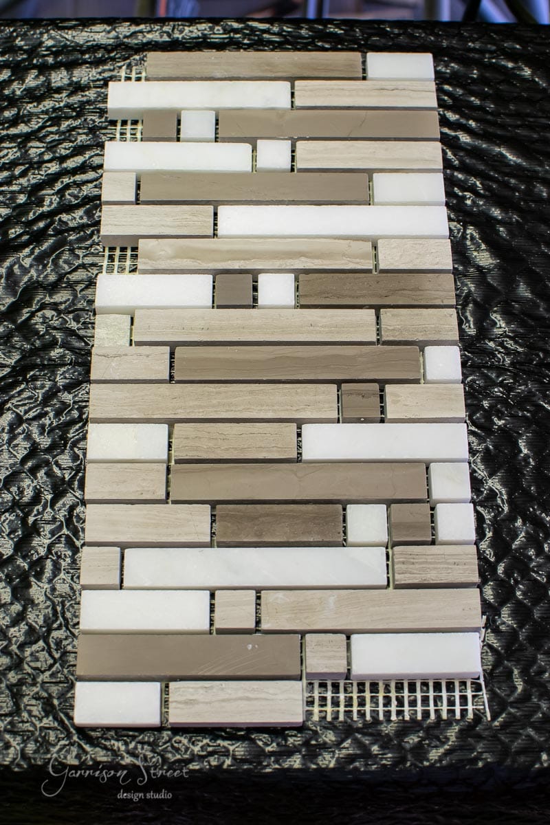 A rectangular tile mosaic displays a blend of beige, white, and taupe tiles in a linear pattern, gracefully set against a textured dark surface.