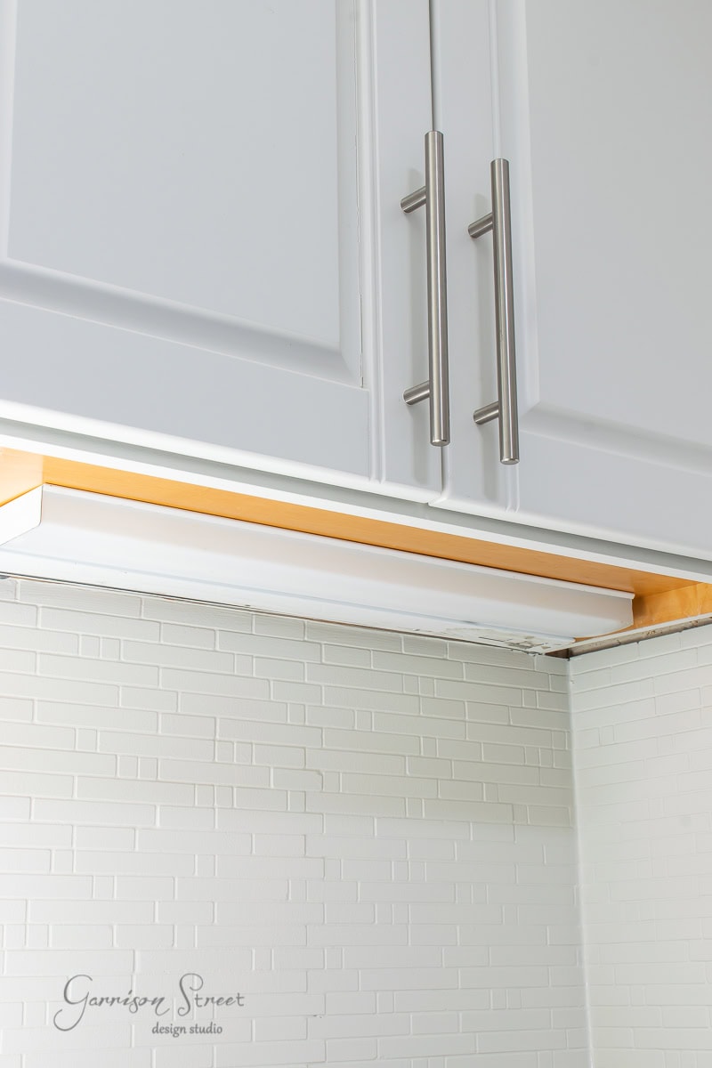 Underneath a white kitchen cabinet, a sleek lighting fixture highlights the light gray tile backsplash with its subtle brick pattern. The cabinet's silver handles add a touch of elegance.