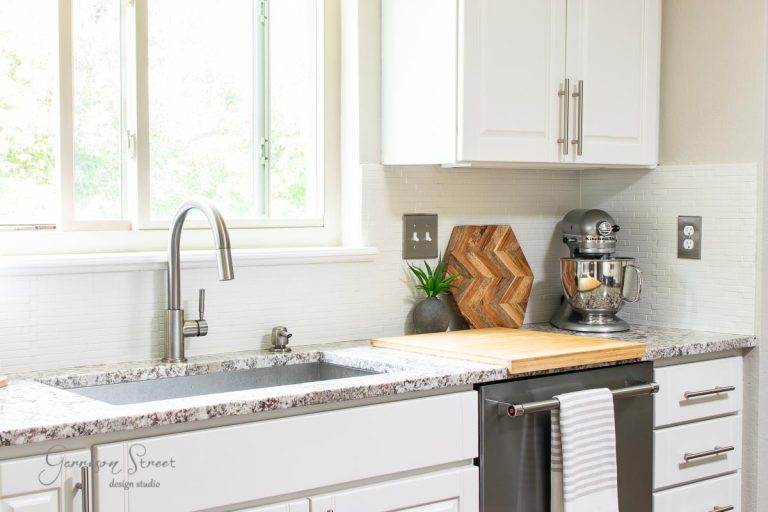 A modern kitchen featuring white cabinets and granite countertops. A stainless steel mixer sits on the counter next to a cutting board, while a window above the sink floods the space with natural light. Amidst this chic setup, a striped towel hangs on the oven handle, adding style tips like how to paint tile.