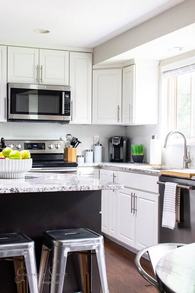 A modern kitchen with white cabinets, stainless steel appliances, and a granite countertop. A bowl of green apples sits on the island with two metal stools. Natural light streams through a window above the sink, where a coffee maker rests nearby.