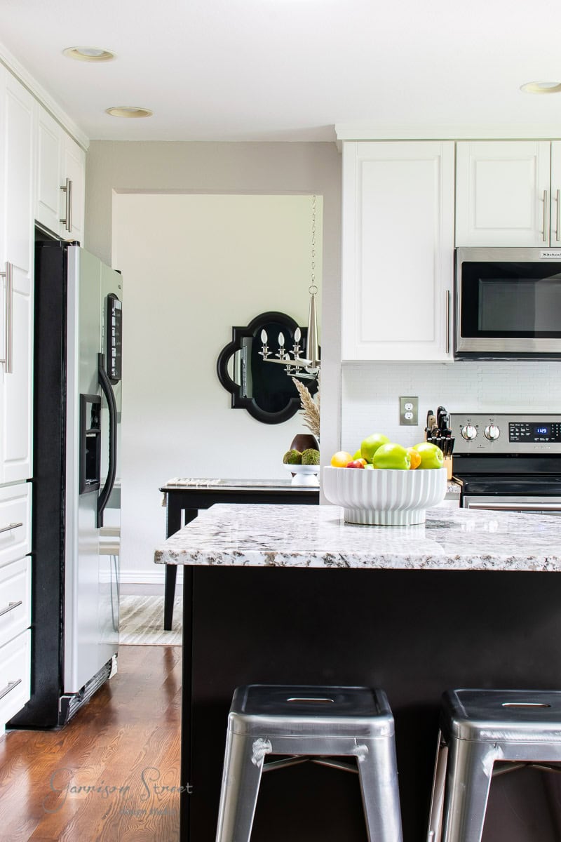 Modern kitchen with white cabinets and a marble countertop island. A bowl of colorful fruit sits on the island, surrounded by metal bar stools. Stainless steel appliances and a chandelier enhance the sleek design.