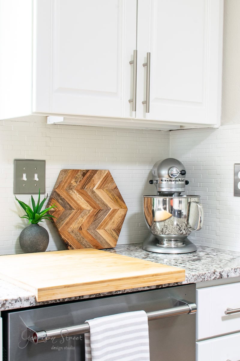 A modern kitchen counter features a gray stand mixer, wooden chevron-patterned board, potted plant, and cutting board. The granite countertop and white cabinets shine. A striped towel hangs from the oven handle.