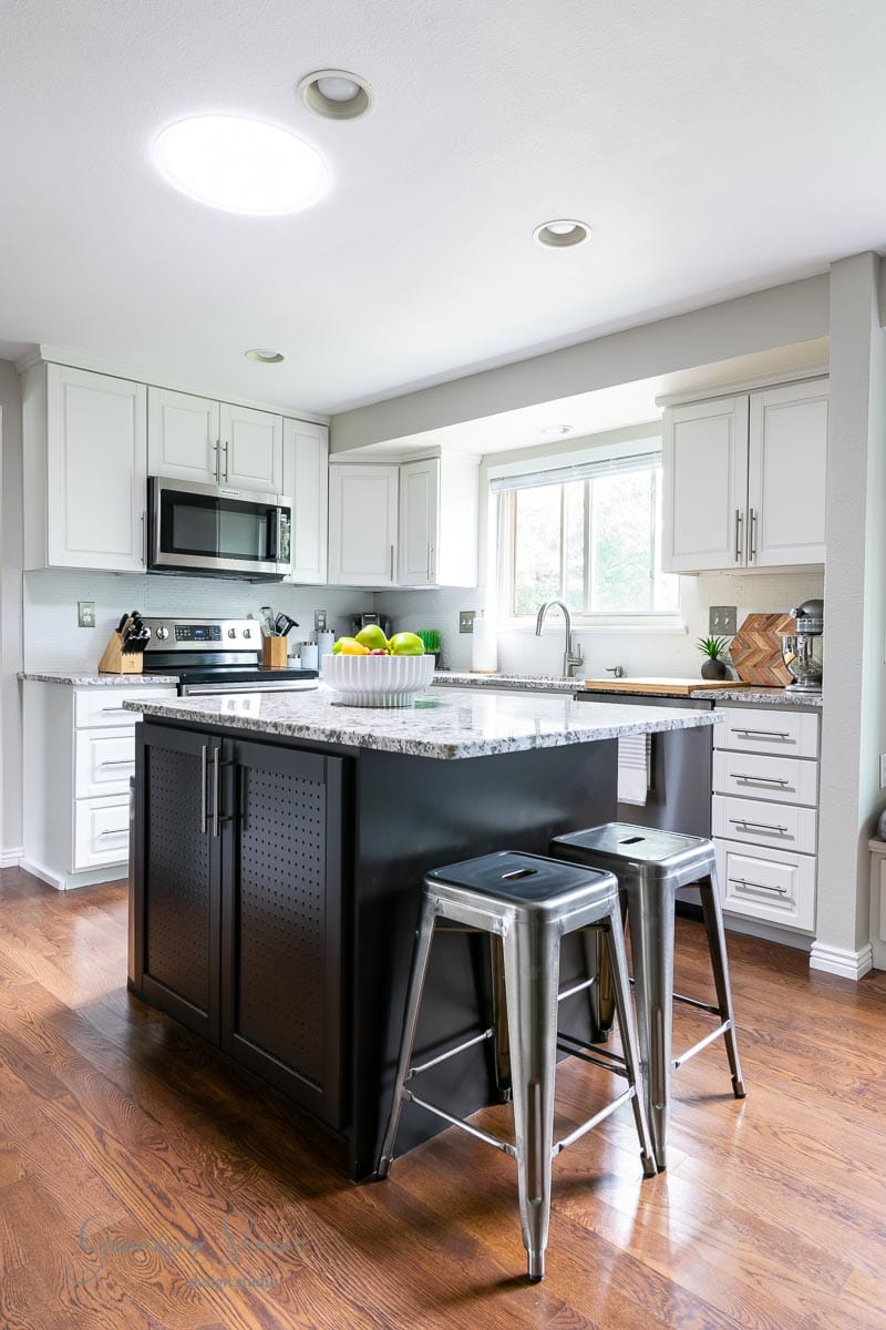 A modern kitchen with white cabinets, stainless steel appliances, and a dark island featuring a speckled countertop. Two metal stools are at the island, with a bowl of fruit displayed. Wooden flooring complements the bright and airy space.