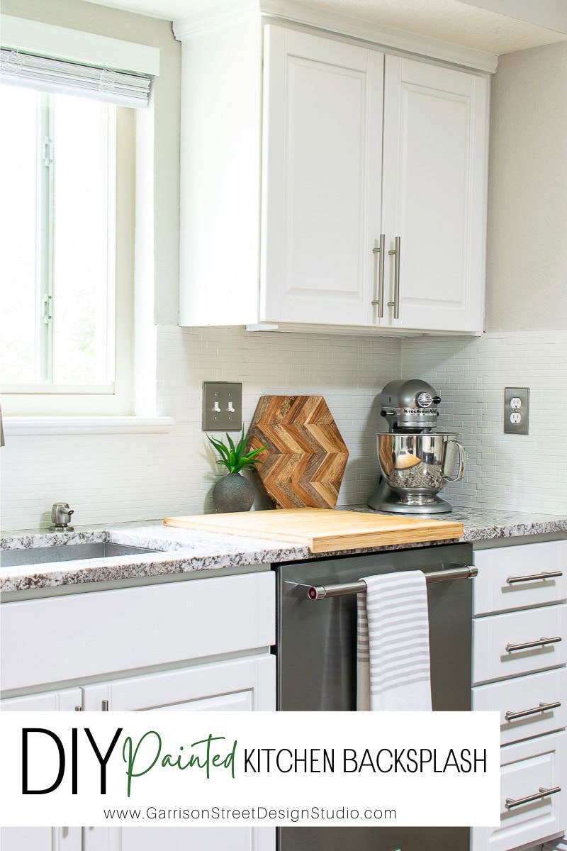 A modern kitchen features white cabinets and drawers, a sleek gray solid surface countertop, and a silver stand mixer. A small wooden cutting board and plant add warmth to the space while the backsplash is elegantly painted white.