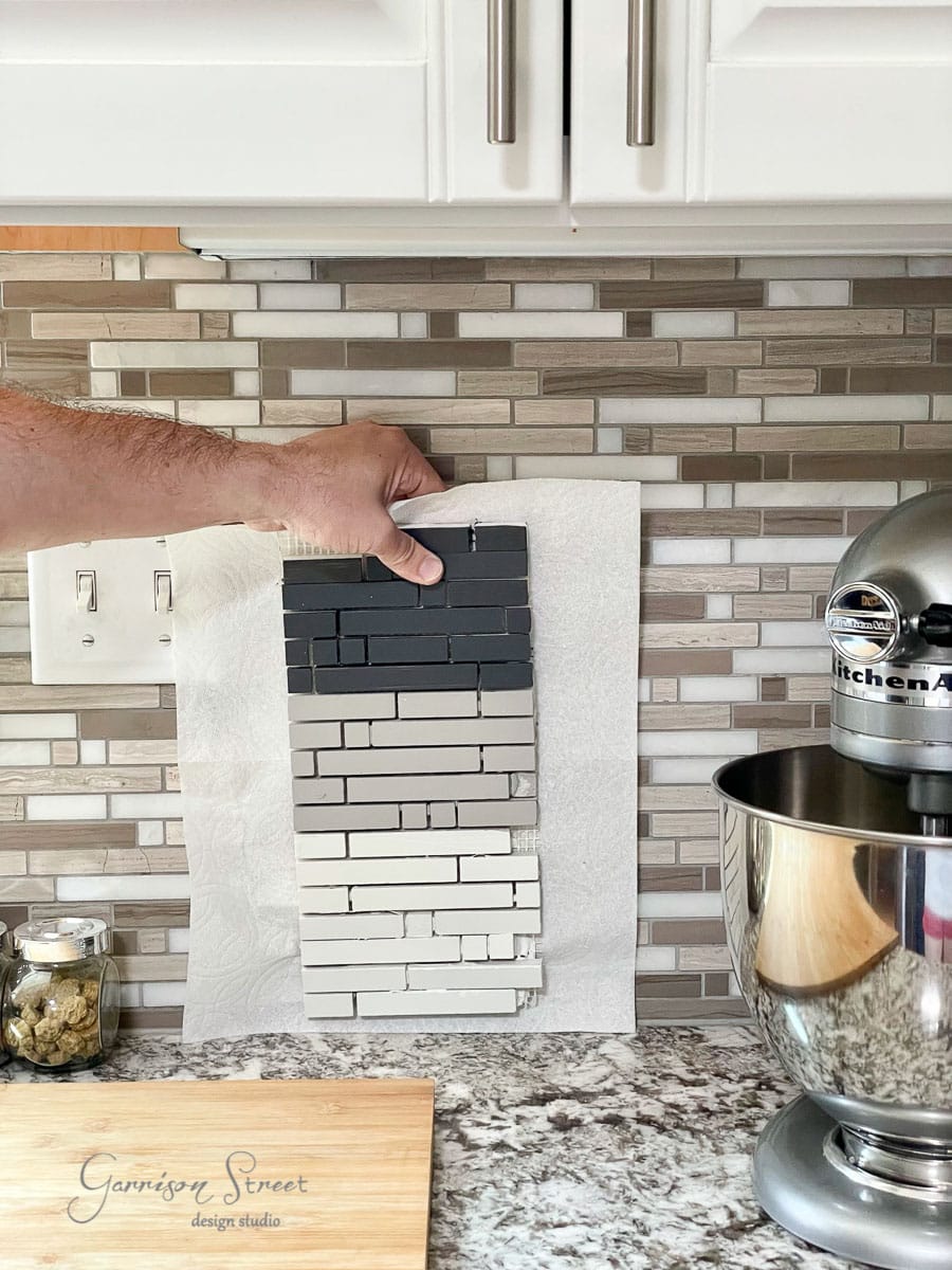 A hand holds a sample of stacked gray tiles against a beige and gray tiled kitchen backsplash. A stand mixer is visible on the right, with a countertop featuring a cutting board and jar on the left.