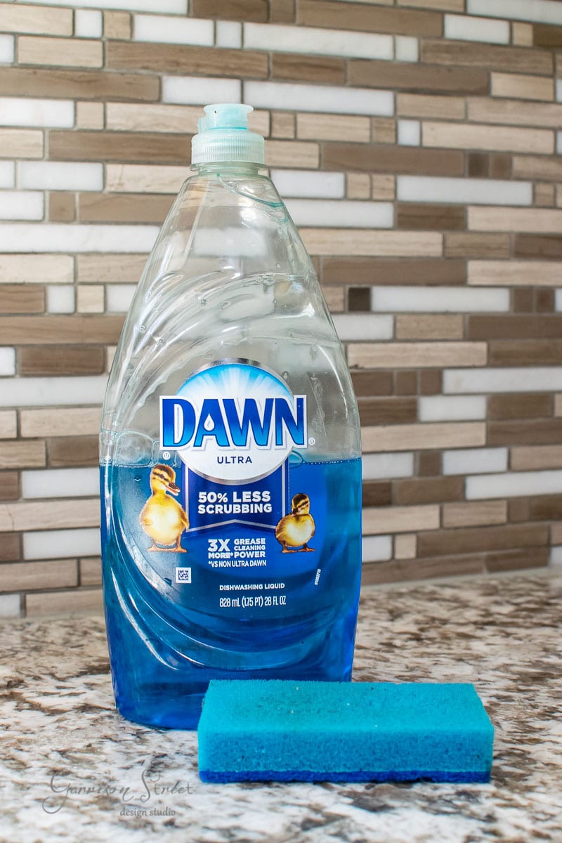 A bottle of Dawn Ultra dish soap with a blue sponge sits on the granite countertop, framed by a mosaic tile backsplash. The label displays a duckling promising "50% less scrubbing."