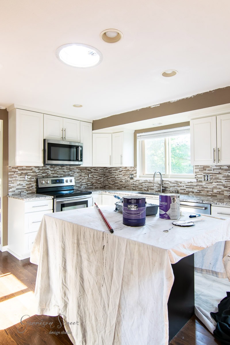A modern kitchen under renovation with white cabinets, a tiled backsplash, and stainless steel appliances. The countertop is covered with a drop cloth. Paint supplies are visible, including a paint can, roller, and brushes. Natural light fills the room.
