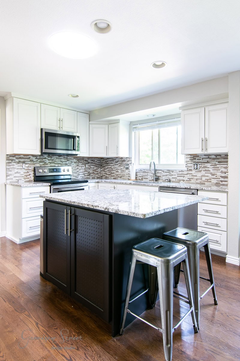 A modern kitchen with white cabinets, a tiled backsplash, and stainless steel appliances. A large island with a granite countertop features two metal stools. The wooden floor complements the natural light streaming through the window.