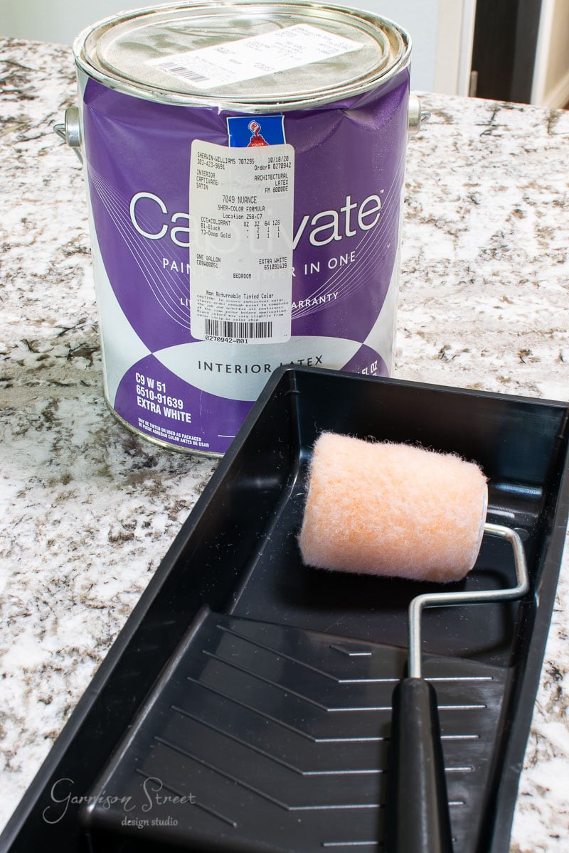 A paint can with a purple label rests on a granite countertop next to a black paint tray and roller brush. A store receipt is taped to the lid, hinting at recent purchases for this creative project.