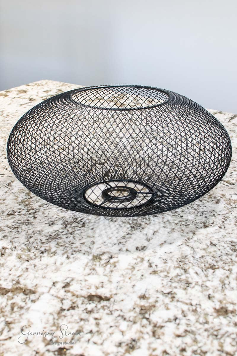 A black, wire mesh decorative bowl from IKEA sits on a granite countertop with a grayish-white pattern. The bowl features an intricate lattice design and is circular in shape, casting a shadow on the surface below.