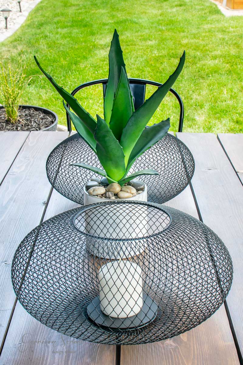 A potted plant with long green leaves sits on a wooden IKEA table. It's surrounded by two wire mesh decorative holders, one containing a white candle, adding to the serene ambiance. The scene overlooks a lush green lawn.