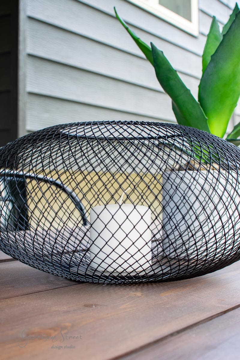 A white candle inside a spherical black metal mesh sits gracefully on an IKEA wooden table. In the background, a large green plant complements the exterior wall of a house with gray siding, creating an inviting atmosphere.