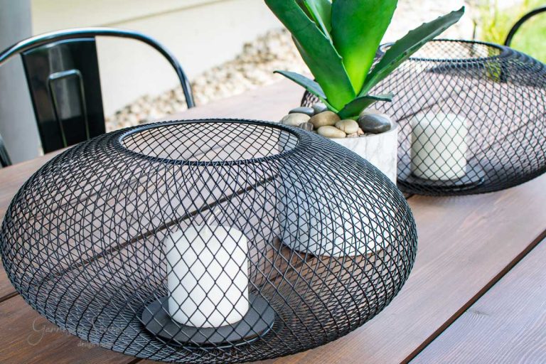 The modern outdoor table setup includes two black wire lanterns with white candles, resembling IKEA's minimalist style. A Luftmassa planter cradles a succulent and stones between them on the wooden table, while a metal chair peeks into the frame.