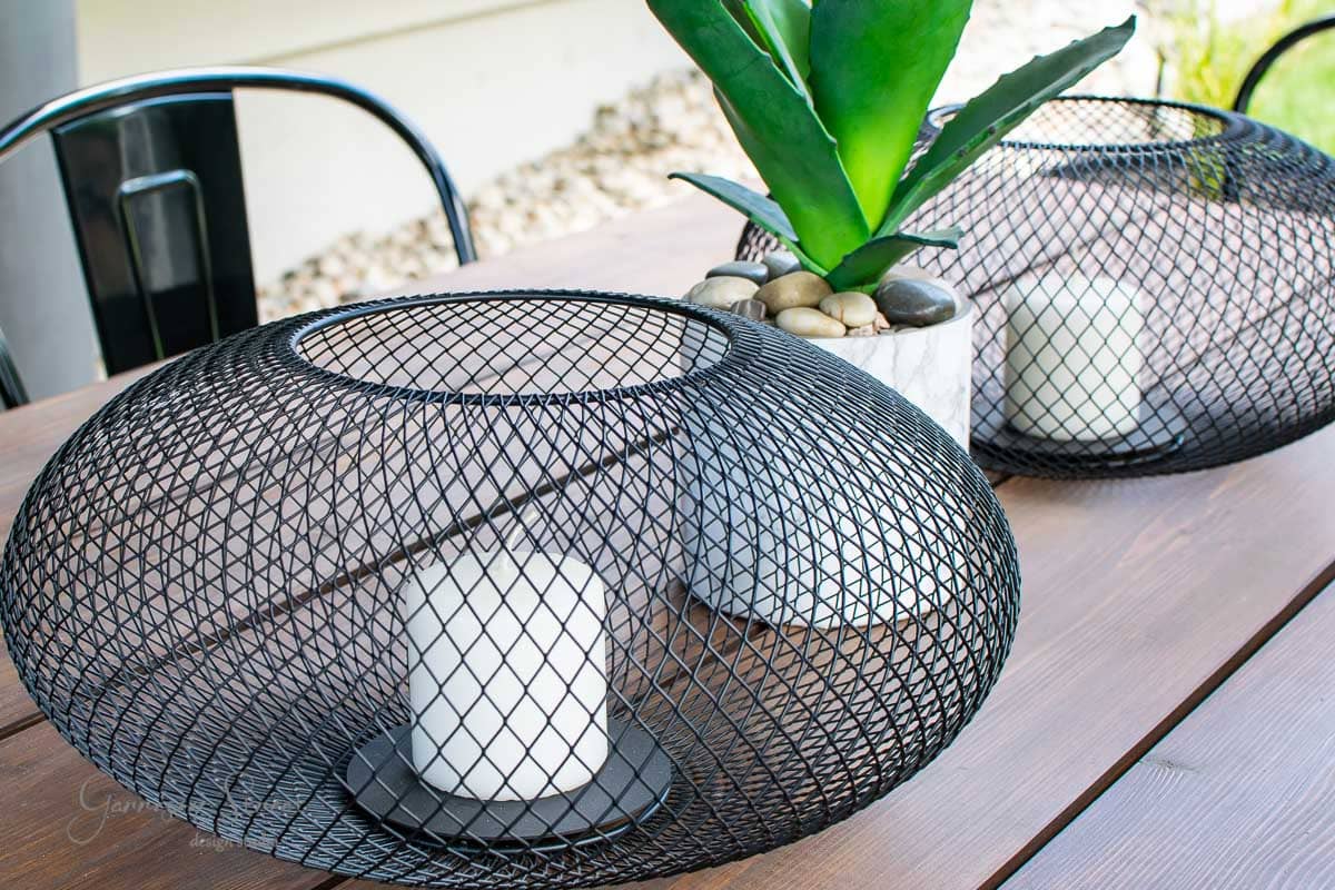 The modern outdoor table setup includes two black wire lanterns with white candles, resembling IKEA's minimalist style. A Luftmassa planter cradles a succulent and stones between them on the wooden table, while a metal chair peeks into the frame.